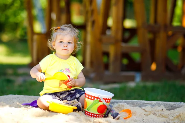 Linda niña jugando en la arena en el patio al aire libre. Hermoso bebé que se divierte en el soleado cálido día soleado de verano. Feliz niño sano con juguetes de arena y en ropa de moda colorida. — Foto de Stock