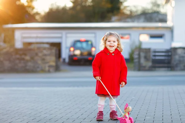 Utomhus porträtt av lilla söta småbarn flickan i röd kappa aon våren solig dag med push träleksak. Friska happy baby barn promenader i staden. Eleganta modekläder för barn. — Stockfoto