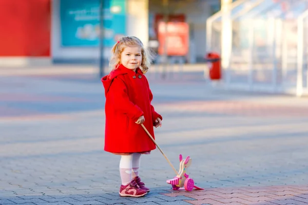 Utomhus porträtt av lilla söta småbarn flickan i röd kappa aon våren solig dag med push träleksak. Friska happy baby barn promenader i staden. Eleganta modekläder för barn. — Stockfoto