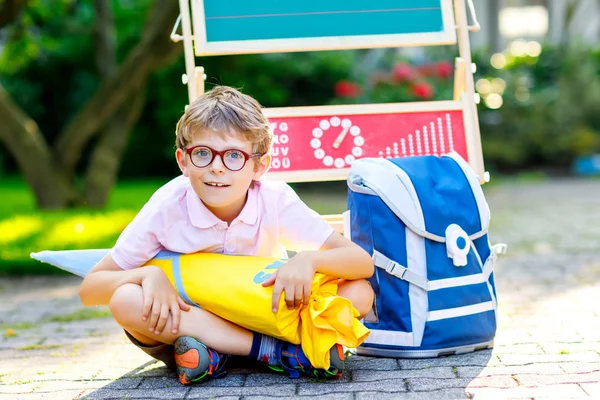 Счастливый маленький мальчик в очках, сидящий за столом, рюкзаком или сумкой. Школьник с традиционным немецким конусом школьной сумки по имени Schultuete в свой первый день в школе — стоковое фото