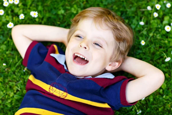 Niño rubio feliz con ojos azules tendido en la hierba con flores de margaritas en el parque. En el cálido día de verano. Niño soñando y sonriendo. Retrato de un niño pequeño o preescolar lindo. — Foto de Stock