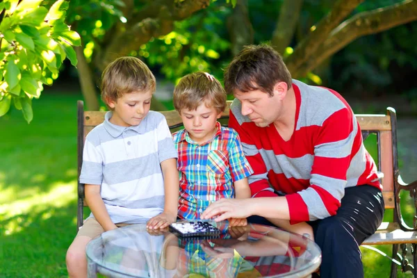 Two funny handsome kid boys and young father playing together checkers game. Sons, siblings children and dad spending leisure together. Family having fun in summer garden outside — Stock Photo, Image