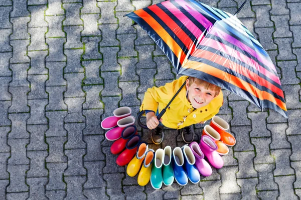 Gyerek kisfiú és a csoport színes eső csizma. Szőke gyermek állandó égisze alatt. — Stock Fotó