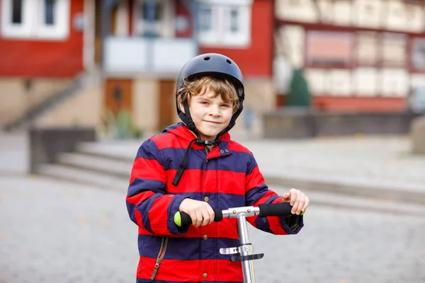 Cute little school kid boy riding on push scooter on the way to or from school. Schoolboy of 7 years driving through rain puddle. funny happy child in colorful fashion clothes and with helmet. — Stock Photo, Image