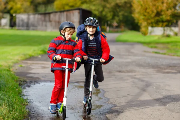 Two little kids boys riding on push scooters on the way to or from school. Schoolboys of 7 years driving through rain puddle. Funny siblings and best friends playing together. Children school out. — Stock Photo, Image