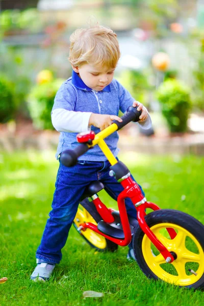 Active blond kid boy in colorful clothes driving balance and learners bike or bicycle in domestic garden. Toddler child dreaming and having fun on warm summer day. outdoors movement game for children — Stock Photo, Image