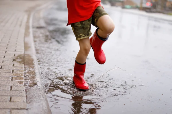 Child wearing red rain boots jumping into a puddle. Close up. Kid having fun with splashing with water. Warm heavy summer rain and happy children — Stock Photo, Image