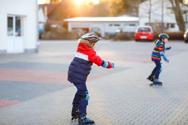 两个小男孩带着滚子在城里溜冰。快乐的孩子，兄弟姐妹和穿着防护服的好朋友。在网上滑板上积极参加体育运动和学习滑冰的男生. — 图库照片