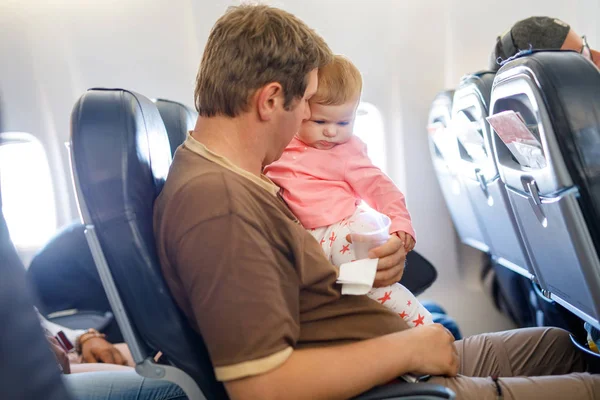 Ung træt far og hans grædende datter under flyvning på fly går på ferie. Far holder og leger med baby pige på armen. Flyrejser med baby, barn og familie koncept - Stock-foto