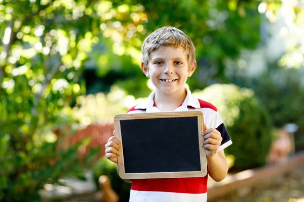 Ragazzino felice con la scrivania di gesso in mano. Sano adorabile bambino all'aperto Scrivania vuota per copyspace tenuta da bella scolara — Foto Stock