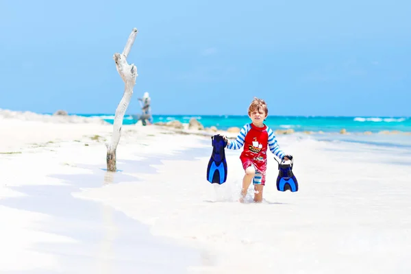 Adorable niño rubio divirtiéndose en la playa tropical de Maldivas. Emocionado niño jugando y surfeando en traje de baño protegido contra el sol en el océano de vacaciones. Arena blanca, niño sosteniendo aletas para nadar —  Fotos de Stock
