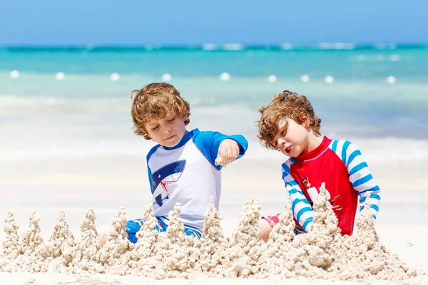 İki küçük çocuk Karayip adasının tropikal sahillerinde kumdan kale inşa ederken eğleniyor. Çocuklar tatillerinde birlikte oynuyorlar. İkizler, mutlu kardeşler gülüyor ve gülüyorlar.. — Stok fotoğraf
