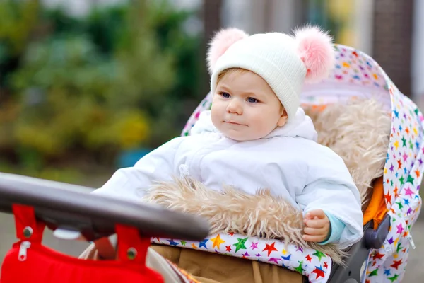 Χαριτωμένο μικρό όμορφο κοριτσάκι κάθεται στο καροτσάκι ή καροτσάκι την ημέρα του φθινοπώρου. Χαρούμενο υγιές παιδί που πάει για περίπατο στον καθαρό αέρα με ζεστά ρούχα. Μωρό με πολύχρωμα ρούχα και καπέλο με μπουμπλς — Φωτογραφία Αρχείου