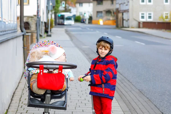 学校への途中でプッシュスクーターに乗ってかわいい小さな学校の少年。ベビーカーや馬車用の車にかわいい赤ちゃん妹と男子生徒。幸せな兄弟. — ストック写真