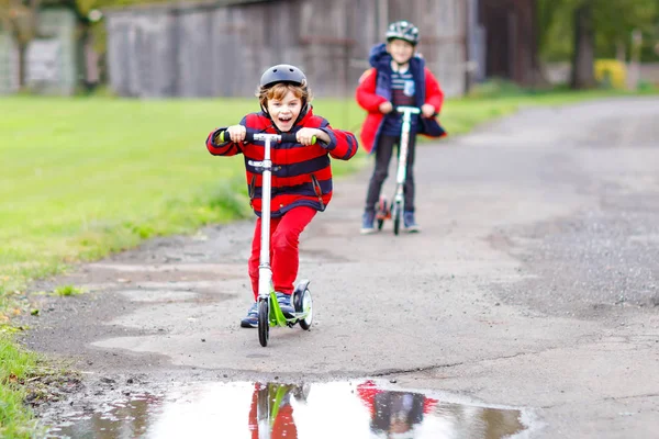 Två små pojkar som åker skoter på väg till eller från skolan. Skolpojkar på 7 år kör genom regnpöl. Roliga syskon och bästa vänner som leker tillsammans. Barn efter skolan — Stockfoto