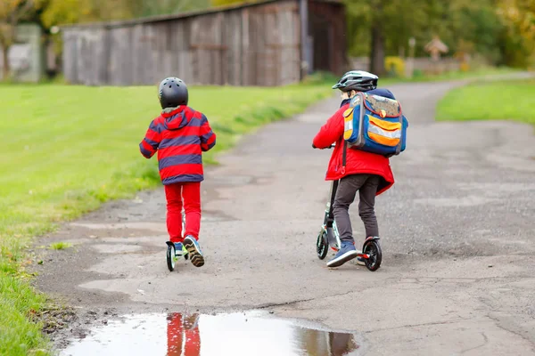 Dos niños pequeños viajando en patinetas de camino a la escuela o desde ella. Colegiales de 7 años conduciendo a través del charco de lluvia. Divertidos hermanos y mejores amigos jugando juntos. Escuela de niños fuera. — Foto de Stock