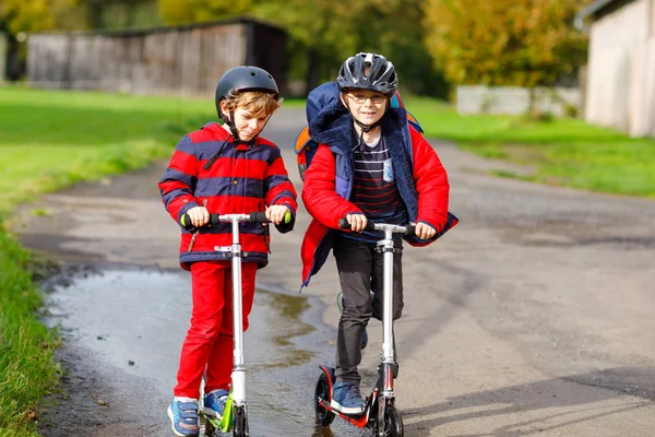 Dos niños pequeños viajando en patinetas de camino a la escuela o desde ella. Colegiales de 7 años conduciendo a través del charco de lluvia. Divertidos hermanos y mejores amigos jugando juntos. Escuela de niños fuera. — Foto de Stock