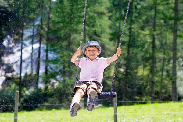 Anak lucu bersenang-senang dengan rantai ayunan di taman bermain luar saat sedang basah memercikkan air. Anak berayun pada hari musim panas. Kesenangan aktif dengan anak-anak. Happy menangis anak laki-laki dengan hujan tetes di wajah. — Stok Foto