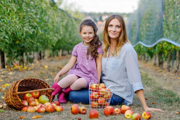 Portrait of little girl and beautiful mother with red apples in organic orchard. Happy woman and kid daughter picking ripe fruits from trees and having fun. Harvest season for family. — Stock Photo, Image