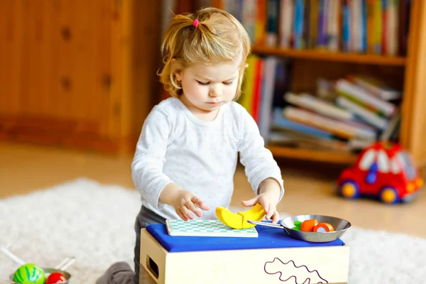 Schattig klein peutermeisje dat thuis speelt met eco houten speelgoed. Gelukkig gezond opgewonden kind snijden groenten en fruit met speelgoed mes. Baby meisje hebben plezier met rol keuken en koken spel. — Stockfoto