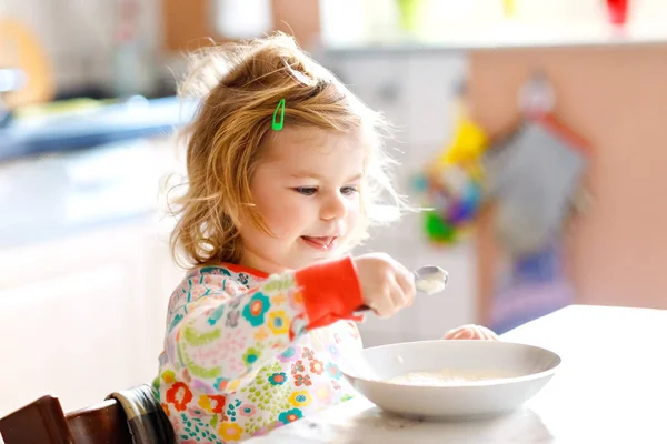 Söt liten flicka som äter hälsosam gröt från sked till frukost. Söt glad baby barn i färgglada pyjamas sitter i köket och lära sig med sked. — Stockfoto