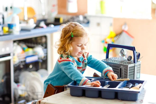 Söt liten småbarn flicka som hjälper i köket med diskmaskin. Glada friska blonda barn sortera knivar, gafflar, skedar, bestick. Baby ha kul med att hjälpa hushållsarbete mor och far. — Stockfoto