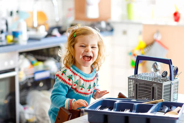 Linda niña pequeña ayudando en la cocina con lavavajillas lavadora. Feliz niño rubio sano clasificando cuchillos, tenedores, cucharas, cubiertos. Bebé divertirse con ayudar a las tareas domésticas madre y padre. —  Fotos de Stock