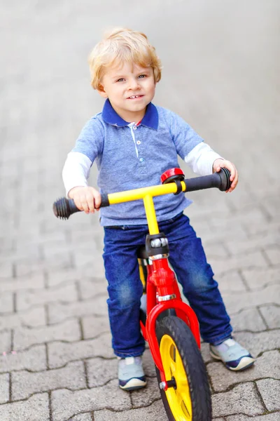 Active blond kid boy in colorful clothes driving balance and learners bike or bicycle in domestic garden. Toddler child dreaming and having fun on warm summer day. outdoors movement game for children — Stock Photo, Image