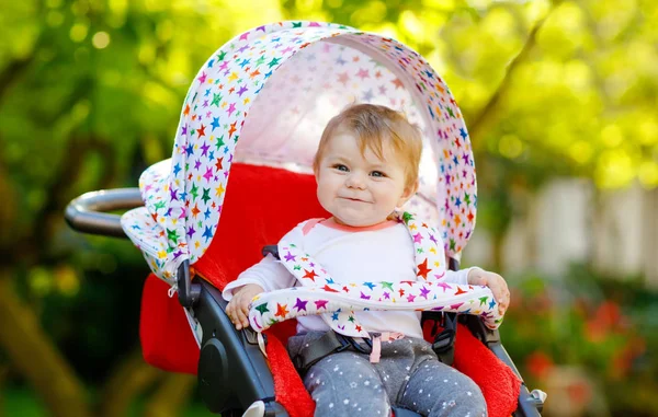 Roztomilé zdravé malé krásné holčičky sedí v kočárku nebo kočárku a čeká na mámu. Šťastné usměvavé dítě s modrýma očima. Se zeleným pozadím. Dcera jde na procházku s rodinou — Stock fotografie