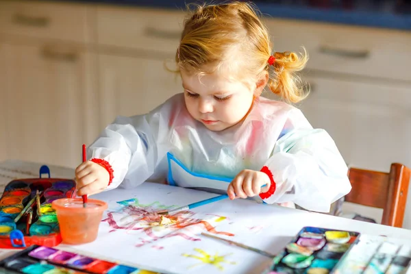 Симпатичная обожаемая девочка учится рисовать акварелью. Маленький ребенок рисует дома, используя красочные кисти. Здоровая счастливая дочь экспериментирует с цветами, водой дома или в детской — стоковое фото