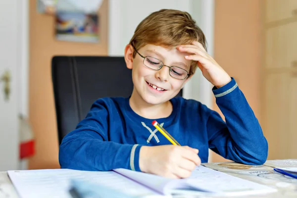 Portrett av en søt skolegutt med briller som gjør hjemmelekser. Liten, konsentrert barneskrift med fargerike blyanter, innendørs. Grunnskole og utdanning – stockfoto