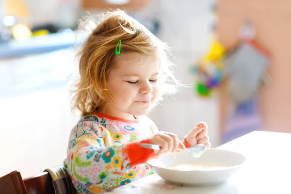 Adorable niña pequeña comiendo porrige saludable de cuchara para el desayuno. Lindo bebé feliz niño en pijama colorido sentado en la cocina y aprendiendo usando cuchara. —  Fotos de Stock