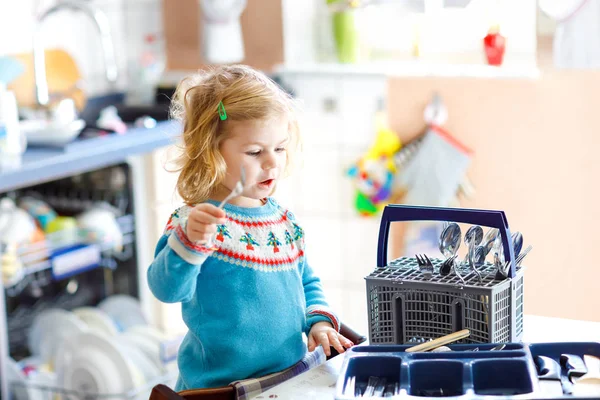 Carina la bambina che aiuta in cucina con la lavastoviglie. Felice bambino biondo sano smistamento coltelli, forchette, cucchiai, posate. Bambino divertirsi con aiutare i lavori domestici madre e padre. — Foto Stock