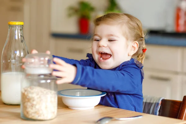 Entzückendes Kleinkind, das gesunde Haferflocken mit Milch zum Frühstück isst. Nettes glückliches Baby in bunten Kleidern sitzt in der Küche und hat Spaß mit der Zubereitung von Hafer, Getreide. Zuhause drinnen — Stockfoto