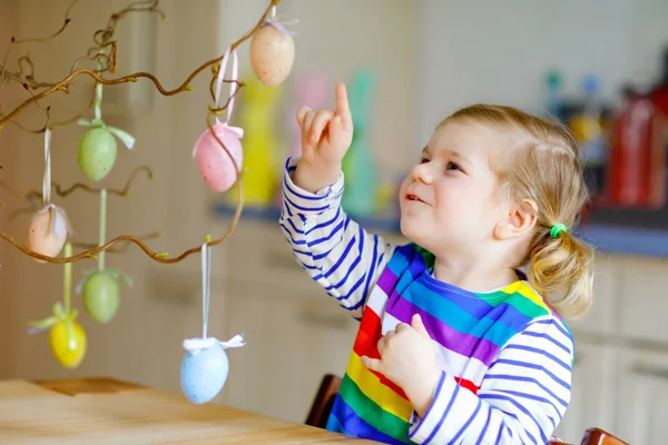 Cute little toddler girl decorating tree bough with colored pastel plastic eggs. Happy baby child having fun with Easter decorations. Adorable healthy smiling kid in enjoying family holiday — Stock Photo, Image