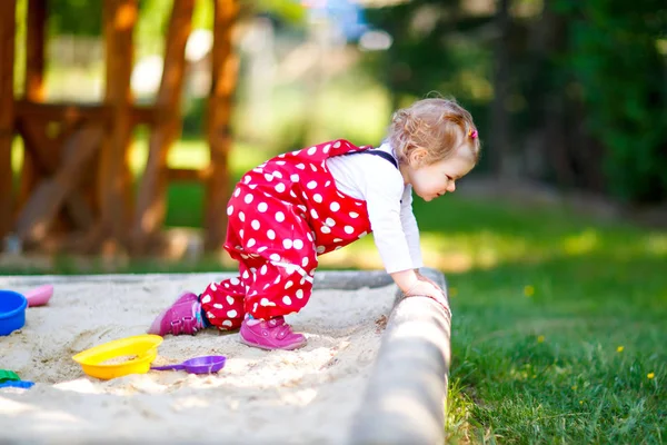 Menina bonito brincando na areia no parque infantil ao ar livre. Bebê bonito em calças de goma vermelha se divertindo no dia de verão quente ensolarado. Criança com brinquedos coloridos de areia. Bebê ativo saudável ao ar livre joga jogos — Fotografia de Stock