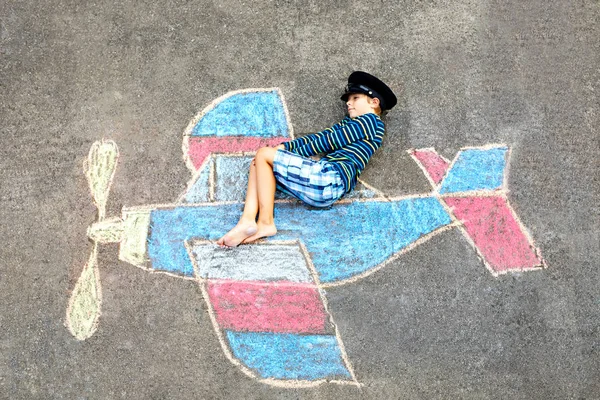 Kleiner Junge, der sich mit Flugzeugbilderzeichnung mit bunten Kreiden auf Asphalt vergnügt. Kindermalerei mit Kreide und Buntstift und Urlaub machen oder vom Pilotenberuf träumen. — Stockfoto