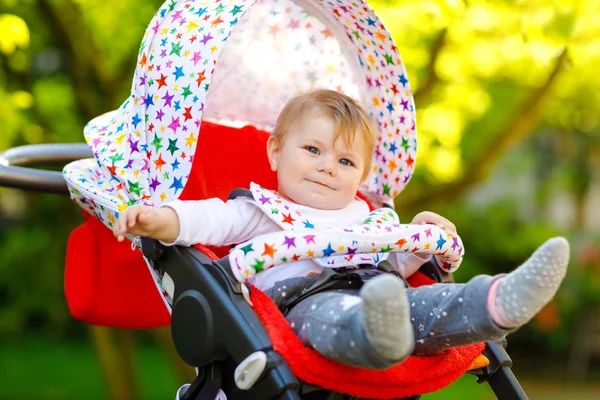 Leuke gezonde kleine mooie baby meisje zitten in de kinderwagen of kinderwagen en wachten op moeder. Gelukkig lachend kind met blauwe ogen. Met groene boomachtergrond. Kleine dochter gaat wandelen met familie — Stockfoto