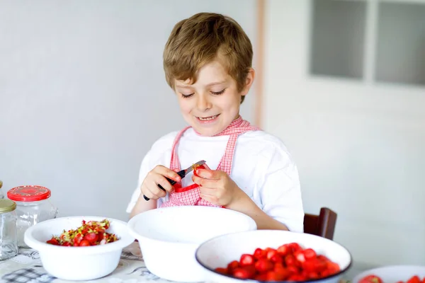 Little blond kid boy helping and making strawberry jam in summer. Funny child cleaning berries and preparing for cooking jam. Kid eating ripe strawberries in domestic kitchen. — Stock Photo, Image