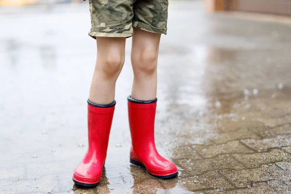 Kind met rode regenlaarzen die in een plas springen. Sluit maar af. Hij heeft plezier met spetteren met water. Warme zware zomerregen en blije kinderen. — Stockfoto