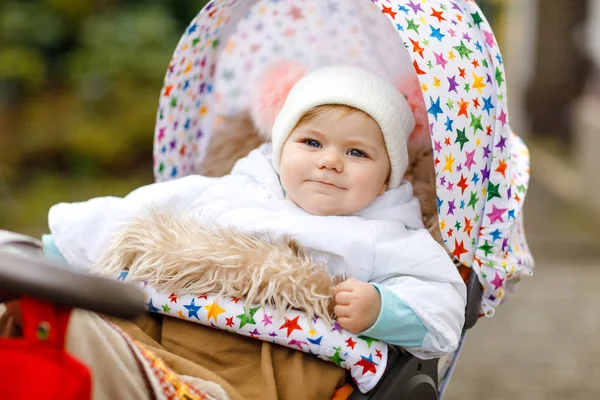 Söt liten vacker flicka som sitter i barn vagnen eller sittvagn på höst dagen. Lyckligt friskt barn går på en promenad på frisk luft i varma kläder. Baby med i färgglada kläder och mössa med Bobbles — Stockfoto