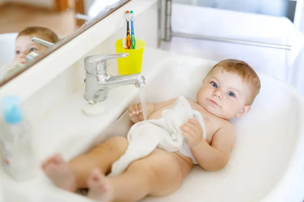 Schattige schattige baby nemen bad in wastafel en pak waterkraan. Klein gezond meisje met grote blauwe ogen die plezier hebben. Schoon mooi kind spelen — Stockfoto