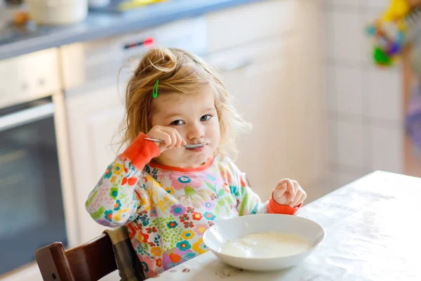 Söt liten flicka som äter hälsosam gröt från sked till frukost. Söt glad baby barn i färgglada pyjamas sitter i köket och lära sig med sked. — Stockfoto