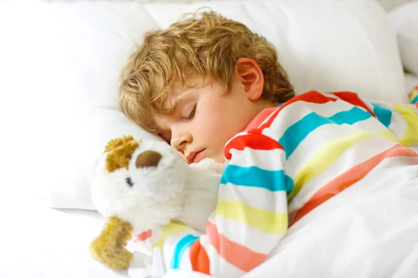 Adorable niño rubio en ropa de dormir colorida durmiendo y soñando en su cama blanca con juguete. niño sano con juguete suave, sueño tranquilo en casa — Foto de Stock