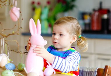 Ağaç süsleyen küçük şirin bir kız ve renkli pastel plastik yumurtalarla tavşan. Mutlu bebek Paskalya süslemeleriyle eğleniyor. Aile tatilinde gülümseyen sevimli çocuk.