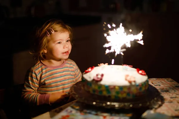 En förtjusande liten flicka som firar sin andra födelsedag. Baby barn äter marshmellows dekoration på hembakad kaka, inomhus. Glad frisk småbarn är förvånad över fyrverkeri glitter på tårta — Stockfoto