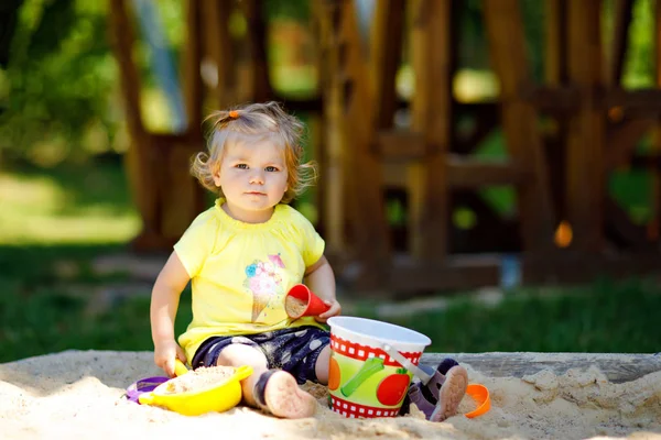 Schattig peuter meisje spelen in zand op buiten speeltuin. Mooie baby die plezier heeft op zonnige warme zomerse zonnige dag. Gelukkig gezond kind met zand speelgoed en in kleurrijke mode kleding. — Stockfoto