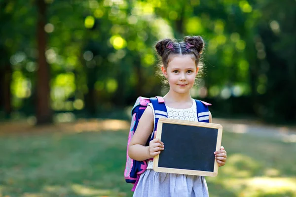 Lycklig liten unge flicka stående med skrivbord och ryggsäck eller skolväska. Schoolkid på första dagen i elementär klass. Friska bedårande barn utomhus, i Green Park. Copyspace tom skrivbord — Stockfoto