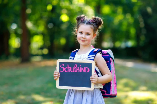 Niña feliz de pie con escritorio y mochila o mochila. Colegial en el primer día de clase elemental. Niño sano y adorable al aire libre, en el parque verde. Sobre el escritorio - Escolar - en alemán — Foto de Stock