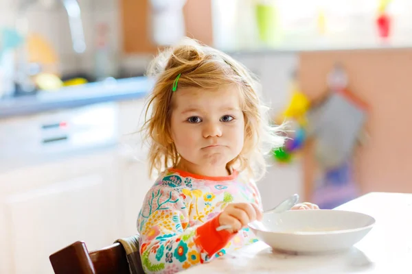 Entzückendes Kleinkind-Mädchen, das gesunde Schweinefleisch vom Löffel zum Frühstück isst. Nettes glückliches Baby im bunten Pyjama sitzt in der Küche und lernt mit dem Löffel. — Stockfoto